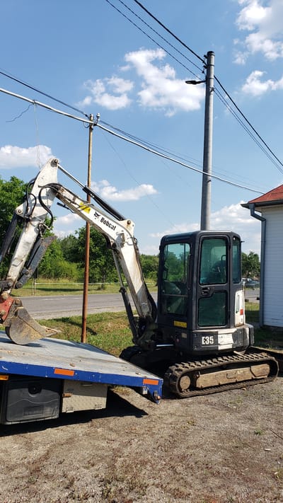 This media file captures a compact excavator, identified as a Bobcat E35, positioned on a flatbed trailer. The excavator is primarily white with black accents, featuring a visible "Bobcat" logo on its arm. The cab of the excavator is dark and tinted. The trailer itself is blue with orange reflectors and appears to be parked on a gravel surface. Behind the trailer, there is a grassy area with trees and a road in the distance. Overhead, a utility pole with multiple wires stretches across a bright blue sky dotted with white, fluffy clouds, suggesting a sunny day. The image is oriented vertically, with the excavator and trailer appearing to be on the right side of the frame. The scene has a functional, industrial feel, likely indicative of construction or landscaping work. The presence of the utility pole and the overcast sky suggest an outdoor setting, potentially in a rural or suburban area. The time of day appears to be daytime, given the bright sunlight. There is no visible activity or people in the frame.