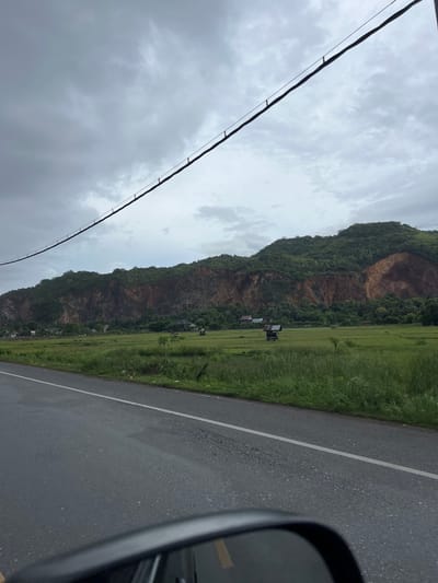 The image depicts a landscape scene in Lampisang, Indonesia, likely captured from a moving vehicle. A dark, asphalt road with a white dividing line occupies the right side of the frame. Adjacent to the road is a lush green field, possibly rice paddies, stretching towards a dramatic, tree-covered mountain range. The mountain face is rugged, with exposed rocky areas contrasting with dense vegetation. A few small structures, possibly sheds or simple houses, are visible nestled amongst the trees and fields at the base of the mountain.

Dominating the left side of the image is a dark power line that stretches diagonally across the frame, supported by visible insulators. Above the landscape, a cloudy, overcast sky fills the upper portion of the image, suggesting it is either early morning or late afternoon, or that the weather is generally cloudy. The overall mood is serene and natural, with the imposing mountain and vast sky creating a sense of scale. No people are directly visible, but the presence of the road and the power line indicates human activity and infrastructure within the natural setting. The image is oriented vertically, with the horizon running across the middle of the frame.