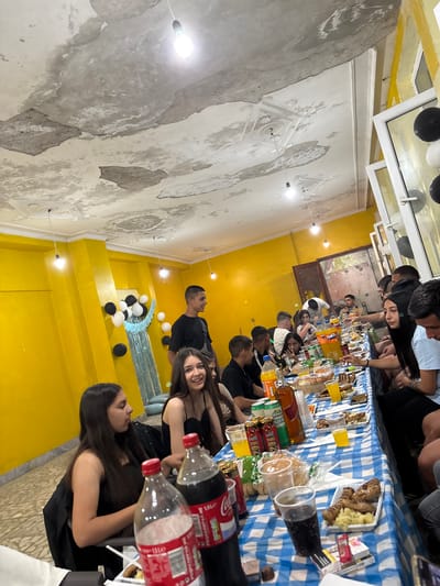 This image depicts an indoor gathering at a venue in Mihailovo, Bulgaria, possibly a celebration or party. A long table is set up, covered with a blue and white checkered tablecloth, laden with food and drinks. Numerous individuals, predominantly young adults, are seated around the table, engaged in conversation and partaking in the meal. Several bottles of soda, including Coca-Cola and Fanta, are visible, alongside glasses and various food dishes, including what appears to be grilled meats and mashed potatoes. Balloons, black and white, are strategically placed as decorations, suggesting a festive occasion. The room's interior features bright yellow walls and a distressed, textured ceiling with exposed light fixtures, indicating a possibly older or utilitarian building. The overall atmosphere appears casual and social, with people interacting and enjoying each other's company.