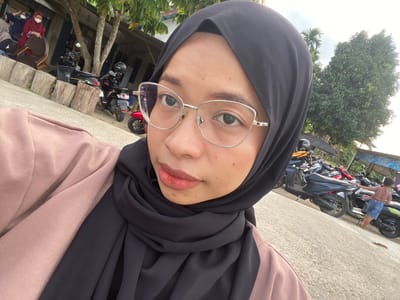 The image shows a young woman wearing a black hijab and glasses, taking a selfie. She is wearing a pale pink sweater or cardigan. In the background, there are several parked motorcycles. To the left, there are some wooden posts and a building with some people. To the right, a young boy in a brown t-shirt and blue shorts is standing near a motorcycle. The scene appears to be outdoors, with trees and a cloudy sky. The location is identified as Air Molek, Indonesia.
