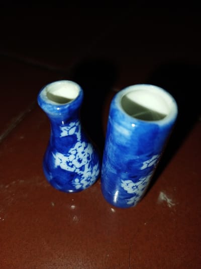 The scene, located in Longmiang, Laos, displays two small, blue and white ceramic objects positioned on a reddish-brown surface. The object on the left is a miniature ceramic container with a bulbous base, a narrow neck, and a subtly flared rim. Its exterior features a dark blue glaze with white, intricate floral or leaf patterns. The internal surface visible through the opening is white. To its right, a slightly taller and more slender cylindrical ceramic container stands upright, exhibiting a similar dark blue glaze with white floral or leaf patterns. The interior of its opening is also white. Both objects are static and closely spaced on the flat, reddish-brown surface. The environment is dimly lit, resulting in a dark upper background and subtle shadows around the objects. Minor surface imperfections are visible on the supporting surface.