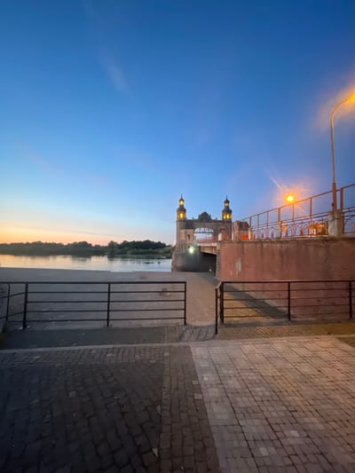 The image shows the Sovetsk Bridge, also known as the King's Bridge, over the Neman River in Sovetsk, Russia, at dusk. The bridge is an imposing stone structure with two towers and a large archway. It is illuminated by lights, giving it a warm glow against the twilight sky. The river is calm and reflects the light from the bridge and the sky. In the foreground, there is a paved walkway with a black metal railing. The sky is a deep blue, with hints of orange and pink from the setting sun on the horizon. The overall impression is one of serene beauty and historical grandeur.