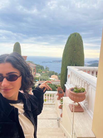 A person, likely female, is taking a selfie on a balcony or terrace in Èze, France. They have dark, curly hair, wear sunglasses, a dark jacket, and a white top with some illegible text. Their left hand is raised, making a two-finger gesture, and a gold ring is visible.

The immediate foreground includes a white balustrade with classic architectural details and a terracotta planter holding small plants. A tall, slender cypress tree stands prominently to the left, partially obscuring the view.

The background reveals a stunning panoramic vista of the Mediterranean Sea or a large bay. Lush, tree-covered hillsides with scattered buildings slope down to the water's edge, and distant landmasses are visible across the calm sea. The sky is partly cloudy, with softer, possibly golden light appearing towards the horizon, suggesting late afternoon or early evening. The person has a slight smile, indicating a relaxed and content mood amidst the beautiful coastal scenery.