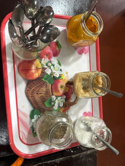 Four small jars with spoons sit on an apple-themed tray alongside a glass of additional spoons. The jars contain turmeric, brown sugar, white sugar, and pepper. A small woven basket with apples and apple blossoms decorates the tray's center. The tray has a white background with a red trim. It rests on a dark surface.