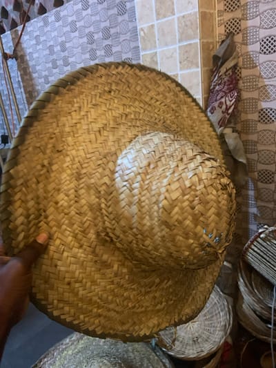 The image displays a large, wide-brimmed straw hat from an overhead perspective, with a person's hand touching its brim. The hat is made of natural, light-colored straw, predominantly golden-yellow and beige, with some darker brownish-greenish tones in places. Its surface exhibits a distinct diagonal basket weave pattern (twill weave) across both the rounded crown and the expansive, flat brim. The crown shows some minor irregularities and slight gaps in the weave near the top, indicating its handmade nature. The edge of the brim is reinforced with a slightly darker, possibly braided, finish.
In the background, a variety of textures and patterns are visible. To the left, the surface beneath or beside the hat appears to be tiled. There are two distinct tile patterns: one section features small, light-colored square tiles in shades of beige and off-white, while another section displays a repeating geometric pattern of interlocking loops or ovals in light gray and white. Above these tiles, a piece of patterned fabric or textile, possibly folded, is visible, featuring a reddish or maroon design on a lighter background.
To the right and in the upper background, several other woven items are stacked or displayed. These items, likely other hats or baskets, are also made of natural fibers and exhibit various weaving patterns and shades of brown and beige, suggesting a setting like a market stall or craft shop. One appears to be a round, flat woven item, possibly a plate or tray, while another looks like a cylindrical object made of vertically arranged reeds or sticks.
A person's right hand, with dark skin, is visible on the lower right side of the hat, with the index finger gently resting on or touching the brim. This suggests interaction with the hat, perhaps examining its texture or quality, or holding it. The lighting is soft and diffused, highlighting the texture of the straw and the surrounding woven goods. No text is visible in the image.