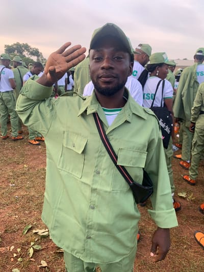 The image portrays a male individual in a light green uniform, performing a casual salute, in an outdoor setting identified as Kubi A, Nigeria. The primary subject, a dark-skinned male with a short beard and mustache, is positioned centrally and faces the viewer directly. He wears a light green collared shirt with two buttoned chest pockets, matching light green trousers, and a baseball cap. A white and green striped undershirt is visible at his neck, and a black cross-body bag with a red and black striped strap is worn across his chest. His right hand is raised near his forehead in a relaxed salute gesture, palm open. In the background, numerous other individuals are visible, largely blurred, also dressed in similar light green uniforms and caps. Some background figures wear white t-shirts bearing the "NYSC" acronym, and several are seen wearing bright orange sandals. The ground is characterized by reddish-brown soil with sparse dry grass and scattered leaves. The sky is overcast, and trees are visible in the distant background.