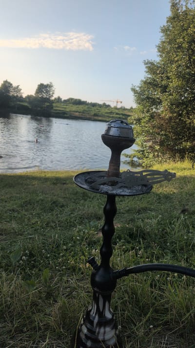 The image shows a close-up view of a hookah resting on the grass near a body of water. The hookah is black with a decorative patterned base. In the background, a body of water is visible, with trees and greenery lining its banks. The sky is clear and blue, with some wispy clouds. A construction crane is visible in the distance. The scene appears to be set outdoors in a natural environment. The lighting suggests it is either early morning or late afternoon. No people are visible in the image. There is no visible text in the image.