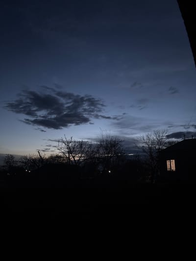 The image shows a night view in Meghrashen, Armenia. The left half of the image depicts a cloudy sky with varying shades of grey and dark blue. A distinct dark cloud formation is visible. The right half is mostly dark, obscuring details. Silhouettes of trees are visible against the fading light of the sky. A building is visible with a lighted window. Additional lights, possibly streetlights, are scattered in the distance.