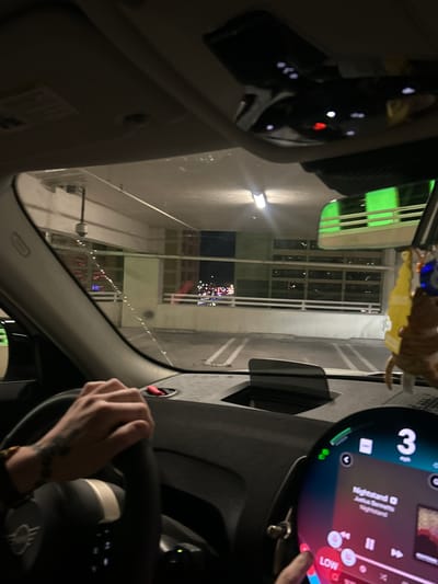 The image depicts the interior of a MINI vehicle at night, positioned within a multi-story parking garage in Reno, Nevada, likely associated with the Peppermill Resort Spa Casino due to the prominent green illuminated building visible in the background and reflected in the rearview mirror.

A person's right hand, displaying a tattoo on the forearm/wrist and wearing a beaded bracelet, grips the steering wheel. A finger from a different hand is interacting with the circular digital infotainment display, which shows "Nightstand" by "Joshua Bassett" playing, a "LOW" indicator, and a speed of "3 mph." Above the dashboard, an empty slot for a heads-up display is visible. From the rearview mirror assembly, a brown, cat-like air freshener, a yellow cutout, and a blue evil eye charm hang.

Through the windshield, which has several water streaks, the interior of the concrete parking garage is visible, illuminated by overhead fluorescent lights. A white security camera is mounted from the ceiling. Multiple empty parking spaces with white markings are present. Beyond the garage structure, distant city lights are visible, along with the distinct green lighting of a building, consistent with the Peppermill Resort Spa Casino in Reno.