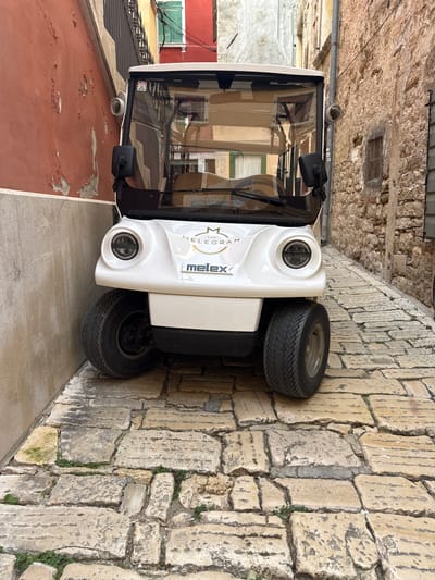 The image shows a white, four-wheeled electric vehicle, specifically a Melex brand, parked in a narrow alleyway in the city of Grad Rovinj, Croatia. The vehicle is oriented facing the viewer, with its front end positioned in the center of the image. The passenger compartment is visible through the windshield. The alleyway walls are constructed of stone. The pavement on the left side of the vehicle is a solid surface, while the pavement on the right is made of stone blocks. The alley appears to be a tight space as the vehicle occupies nearly its entire width. The photograph is taken during the daytime.