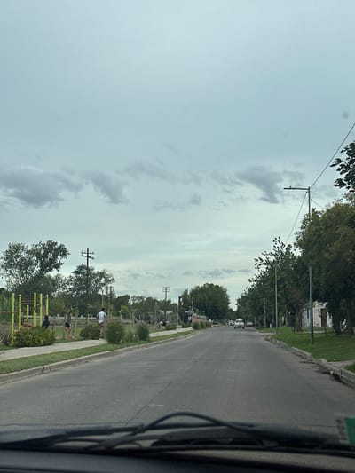 The image depicts an overcast daytime scene on a street in San Miguel, Argentina, viewed from inside a vehicle, evidenced by the dashboard and windshield wipers in the foreground. A two-lane asphalt road extends centrally into the distance, appearing dry.
On the left side of the road, a grassy median separates the roadway from a linear park area. This park contains green lawn, shrubs, and several mature trees. Prominently featured are yellow and green outdoor exercise bars. Four individuals are visible in this park; two are standing near the exercise equipment, and two others are further back, also standing, suggesting recreational activity. Utility poles with wires traverse the scene on this side. Further down the road, additional park features and a large, dark, undefined object are visible.
On the right side of the road, a grassy verge borders a continuous line of mature trees. Streetlights mounted on dark poles are interspersed among the trees. In the distant background on this side, some residential structures are partially obscured by foliage.
Several vehicles are visible on the road in the distance, heading away from the viewer. The sky is largely covered by uniform grey clouds with some lighter patches.