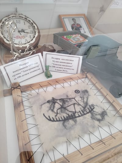 The image displays a glass-enclosed exhibit, likely in a museum or public display area in Yashkino, Russia. The focus is on several items. Prominently featured is a vintage-style analog clock with a decorative floral face. To its right, a framed photograph of a young man in athletic attire is visible. In front of these, there are two rectangular informational plaques. The left plaque, partially obscured, reads in Russian, "Gift from graduates of 1975 group 'Electrician' for the 100th anniversary of the technical school." The right plaque states, "Gift from the collective of the Museum-Reserve 'Tomskaya Pisanitsa' for the 100th anniversary of the technical school, June 2, 2023." A small, green, dragon-shaped figurine sits on the right plaque. Below these, a piece of light-colored fabric, possibly felt, is stretched on a wooden frame with black strings. This fabric features a dark, stylized drawing of what appears to be an animal or mythological creature with intricate patterns. The background is neutral and appears to be a wall. The overall impression is that of a historical display showcasing educational and cultural artifacts.