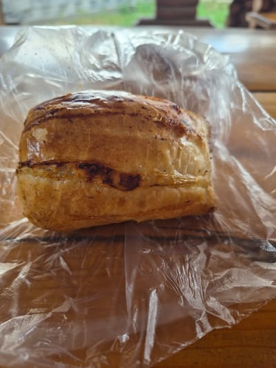 The image shows a close-up of a pastry wrapped in clear plastic, resting on a wooden surface. The pastry appears to be golden brown with some darker, possibly burnt, areas on its crust. The plastic wrap is wrinkled and slightly transparent, allowing some of the background to be visible. The background suggests an outdoor setting, possibly a cafe or restaurant with a blurred view of greenery and wooden structures. No specific landmarks or details pointing to the city of Tekeli, Kazakhstan, are identifiable in the image.