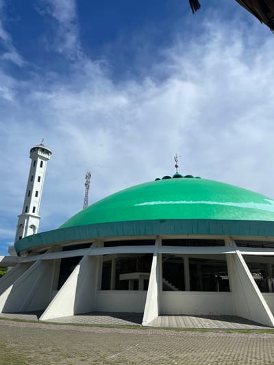 The image shows a mosque in the city of Pasar Paloh, Indonesia. The mosque has a prominent green dome and a tall, white minaret. The building's architecture features a modern design with angular supports for the dome. A telecommunications tower is visible in the background. The sky is blue with scattered clouds. The ground in front of the mosque is paved with interlocking bricks and some grass. There are no people visible in the image.