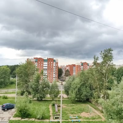 This is an outdoor, eye-level, medium shot of a residential area in Chaikovsky, Russia, on an overcast day. The scene is dominated by large, multi-story brick apartment buildings with green roofs. In the foreground, there is a patch of green grass with trees and bushes. A playground with a slide, swings, and benches is visible. A dark blue car is parked on a paved surface to the left of the frame. In the background, more apartment buildings and trees can be seen, extending to a line of dense forest. The sky is filled with heavy, grey clouds, suggesting gloomy weather. The overall mood is one of quiet residential life under a somber sky. There is no visible text in the image.