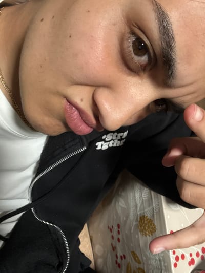A close-up selfie of a young person with light brown skin, brown eyes, and dark eyebrows. The person has a slight frown and pursed lips. There are a few moles visible on their face. They are wearing a white t-shirt and a black zip-up jacket with the words "Str Tghtr" printed on it. A gold chain necklace is visible around their neck. A hand with short fingernails is partially in the frame, with a finger pointing down. The background includes a patterned surface with pine cone and red berry designs. There is no evidence to suggest this picture was taken in Pirdop, Bulgaria.