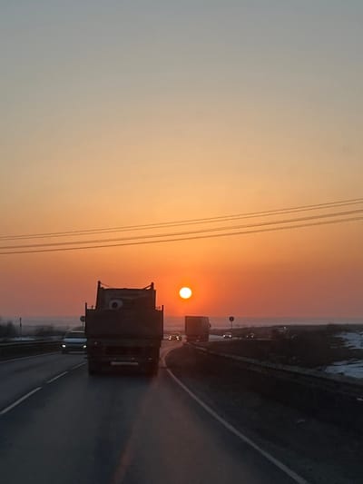 The image depicts a highway scene at either sunrise or sunset in 50 Лет Победы Снт, Russia. The sky dominates the upper portion of the frame, displaying a gradient from pale blue-grey at the top to vibrant orange and red closer to the horizon. A distinct, bright orange sun is positioned low and slightly right of center on the horizon. Four parallel power lines or utility wires traverse the upper middle section of the sky horizontally.
The perspective is from inside a vehicle traveling on a multi-lane asphalt highway. In the immediate foreground, a dark, heavy-duty truck with a high, open cargo bed is directly ahead, facing away from the viewer. Its rear features a rectangular license plate area and unlit taillights. To the left of this truck, in the adjacent lane, a lighter-colored car or SUV is visible with illuminated headlights. Further down the road in the right lane, another dark truck, possibly a semi-trailer, is visible in silhouette. Multiple other vehicles with illuminated headlights appear as distant bright points on the highway.
The road surface is dark and appears slightly reflective, with white lane markings defining the lanes. The highway curves gently to the right in the distance. To the left of the highway, the distant landscape is dark and indistinct, possibly featuring low-lying vegetation or trees. To the right of the highway, the terrain is flatter with visible patches of snow or light-colored ground cover, suggesting a cold season. Several road signs or poles are visible along the right side of the highway. No identifiable individuals are visible in the scene.