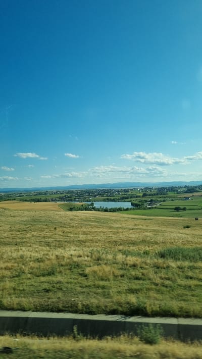 The image captures a wide-angle view of a landscape under a bright blue sky with scattered white clouds. In the foreground, a dry, grassy field transitions to a greener, more manicured lawn. A low, concrete barrier runs along the bottom of the image, suggesting a roadside perspective.

Further in the midground, rolling hills are dotted with trees and what appear to be cultivated fields of varying shades of green and yellow. A body of water, likely a lake or pond, is visible, reflecting the sky. Beyond the water, a town or village is nestled, characterized by numerous buildings with red roofs, interspersed with more greenery. In the distance, a range of mountains forms the horizon. The scene is likely a rural or semi-rural setting, possibly on the outskirts of the city of Bulai, Romania, given the typical landscape of the region. There are no people or animals clearly visible in the image.