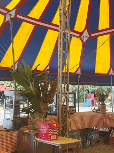 The image depicts the interior of a blue and yellow striped tent, possibly at a fair or event, with a central metal support structure. The foreground features a red and white checkered tablecloth covering a table, with various items such as a popcorn machine, a small fan, a basket, glass jars, and a pink box with white writing that says "Moscato White Wine Valencia". Below the table, several cases of bottled water are visible on wooden pallets. In the background, beyond the tent opening, a playground slide and trees are visible. The overall scene suggests an outdoor event venue. The location is specified as Gueutteville, France.