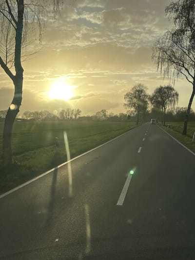 The image captures a rural road in Ottersberg, Germany, at sunset. The road is asphalt, with white lane markings. A vehicle is seen in the distance. Fields flank the road, and bare trees line the horizon and foreground. The sky is a gradient of yellow and white, with visible cloud formations. The sun is low, creating a bright glare. The scene is quiet and serene.