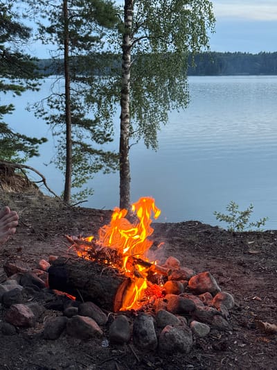 A campfire burns brightly on a rocky shore overlooking a calm, blue lake. The scene is set in a natural environment with trees lining the bank, their branches reaching towards the sky. A human foot is visible in the lower left corner of the frame, close to the fire. The fire is contained within a ring of stones, with logs and branches fueling the flames. The water appears still, reflecting the soft light of the sky. The overall impression is one of a peaceful outdoor setting.