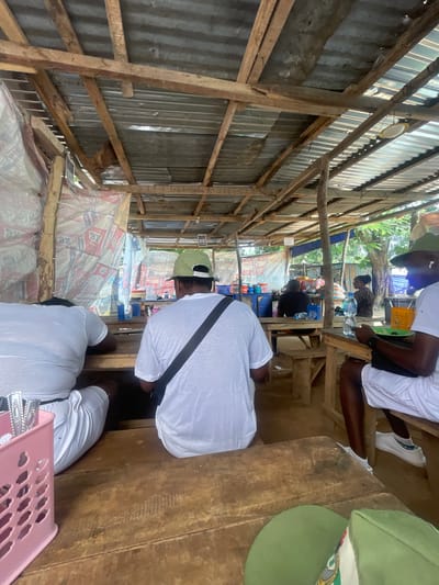 The image depicts an open-air eating establishment under a rustic, wooden-framed roof with a corrugated metal covering. The structure appears to be located in Orun Ekiti, Nigeria. Several individuals are seated at wooden tables, some of whom are wearing white t-shirts and shorts. One person, in the foreground, is wearing a green cap and white t-shirt and shorts. Another person, also in white, is seated further back and to the left. A third person, in a black shirt, is visible further into the establishment.

The tables are simple wooden planks, and some have plastic containers and bottles of water on them. The walls are partially covered with what appear to be large printed banners or tarps. The general ambiance suggests a casual, local eatery. The lighting is natural daylight, filtered through the open sides of the structure and gaps in the roof. The ground appears to be dirt or concrete, typical of an outdoor setting. The overall scene captures a moment of everyday life at this local food stall.