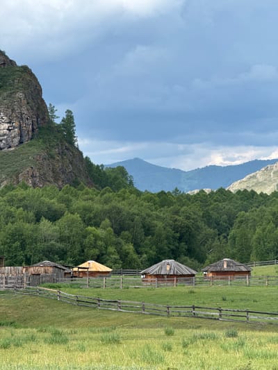 The image depicts a rural scene in the village of Kazanovka, Russia. In the foreground, a wooden fence encloses a grassy field. Behind the fence, several traditional wooden structures are visible, resembling yurts or roundhouses with thatched roofs. One of these structures has a bright yellow, conical roof. A larger wooden building with a sloped roof is situated to the left of these. The middle ground is dominated by a dense forest of green trees. In the background, majestic mountains rise, with the closest ones covered in trees and the further ones displaying rugged, rocky slopes. The sky above is a mix of gray and white clouds. The scene suggests a tranquil, natural landscape with elements of traditional architecture.