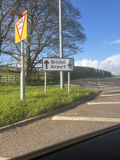 The image displays a roadside scene in Pensford, United Kingdom, under a clear blue sky with scattered white clouds on the horizon. The foreground shows an asphalt road surface with two parallel white markings. A grey concrete kerb separates the road from a lush green grassy verge containing scattered yellow wildflowers.

Prominently positioned on the left side of the verge are two road signs. The first, closer to the viewer, is a yellow triangular "GIVE WAY" sign with a red border and black text, mounted on a grey pole topped with a small black solar panel. Adjacent to it and slightly further back is a white rectangular sign displaying "Bristol Airport" in black text, accompanied by an upward-pointing arrow on the left and a stylized airplane icon on the right. This sign is supported by two grey poles.

Behind the signs and to the left, a large, bare deciduous tree with an extensive branch network stands against the sky. Further left, green fields are visible beyond a wire fence. To the right of the signs, a paved road curves sharply away into the distance, bordered by a dense green hedge. Along this curving road, a tall grey street light pole is visible, along with another smaller triangular warning sign in the distance. The bottom edge of the image features a dark horizontal line, suggesting the interior frame of a vehicle from which the photograph was taken. No individuals or vehicles are present in the observable scene.