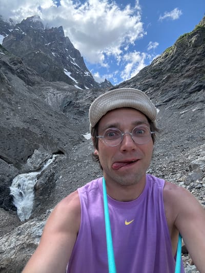 A selfie is taken by a man wearing round glasses, a light-colored corduroy cap, and a purple tank top. He has his tongue sticking out. The man is in a mountainous, rocky environment with a glacier and a waterfall in the background. The sky is blue with scattered white clouds. The location is Murshkeli, Georgia.