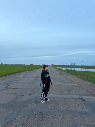 An individual is standing on a paved road that stretches into the distance, with fields and telephone poles on either side. The sky above is overcast. The road exhibits cracking and patching. The person is wearing dark clothing and distinctive black and white sneakers. They are looking upwards. The precise location within Azyakul', Russia is not discernible from the provided image.