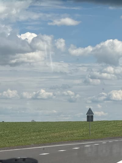 The image shows a rural landscape with a road in the foreground, a green field in the middle ground, and a cloudy sky in the background. A traffic sign, shaped like a black triangle with a white horizontal line across it, stands on a pole in the field. The field appears to be cultivated, with rows of plants visible. In the distance, a lone tree can be seen on the horizon. The sky is filled with a mix of white and gray clouds, with patches of blue sky visible. The road is paved and has white lane markings. The overall impression is one of a quiet, open, and somewhat overcast day in the Belarusian countryside. The scene is devoid of any people or animals. The traffic sign suggests this is a road with specific traffic regulations. The location is identified as Акцябрскі сельскі Савет, Belarus.