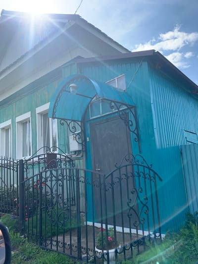 The image shows the exterior of a turquoise-colored wooden house with a dark metal door, situated behind a black wrought-iron fence. The house has several white-framed windows and a blue arched awning over the entrance. A light fixture is attached to the awning. A white electrical box is mounted on the wall next to the door. In front of the house, there is green grass and some potted plants with red and pink flowers. The sun is shining brightly, creating lens flares and highlighting the corrugated metal siding on the right side of the house. The sky above is blue with scattered white clouds. The house appears to be located in a rural or suburban area, possibly in the city of Barsai, Russia, given the context of the request.