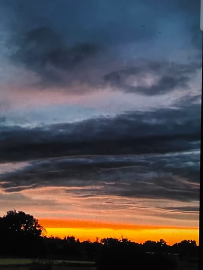 This is a vertical, wide-angle photograph of a dramatic sunset over a rural landscape. The sky is filled with thick, dark clouds in various shades of gray and blue, with a bright band of orange and yellow light emanating from the horizon. The clouds are layered, with some appearing to be illuminated from behind by the setting sun. In the foreground, the silhouettes of trees and a low horizon line are visible. The setting is a rural area, likely on the outskirts of Dubno, Ukraine. There are no people or animals visible in the image. The overall mood is one of dramatic natural beauty.