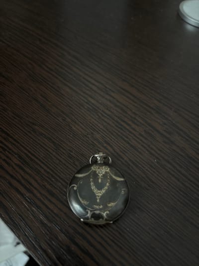 A close-up, slightly angled shot shows a single pocket watch on a dark wooden surface. The watch is round, with intricate decorative patterns on its dark casing, primarily in a lighter, metallic color, possibly gold or silver. The watch has a silver bail loop attached to its top. The wooden surface has a prominent grain pattern, running diagonally from the bottom left to the top right of the frame. The lighting appears to be dim and directional, casting subtle shadows. The background is dark and out of focus, suggesting a close-up perspective. No people or specific locations within Kinel'-Cherkasy, Russia are identifiable in this image.