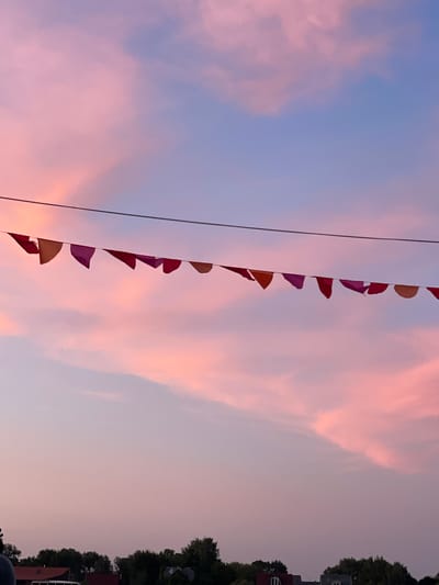 A string of triangular, colorful flags hangs across a pale wire against a soft pink and blue sky. The flags are a mix of orange, pink, and purple hues, and they are spaced out evenly along the wire. In the background, the sky is filled with wispy clouds, tinged with the warm colors of sunset or sunrise. Below the sky, a line of dark green trees and rooftops of houses are visible, suggesting an outdoor scene, possibly in a residential area. The overall impression is peaceful and serene, with a touch of festive decoration provided by the flags.
