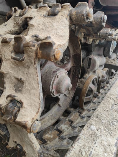 The image shows a close-up, low-angle view of the muddy tracks and wheels of a heavy piece of construction equipment, possibly a bulldozer or tractor. The tracks are made of interlocking metal segments, caked with dry, light-colored dirt and some rust. Several spoked wheels, also covered in dirt, are visible beneath the tracks, some appearing to be idler wheels and others road wheels. One prominent red-painted component, possibly a hub or part of the suspension, is visible towards the center of the frame. The overall impression is one of aged, weathered machinery that has been used in a dusty, outdoor environment. The location is identified as Zashchita, Russia, suggesting an industrial or agricultural setting within that region. There are no people or active processes depicted in the image.