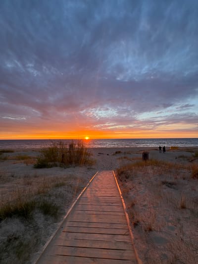 The image depicts a sunset at a beach in Divezeri, Latvia. The sun is setting over the water, with the sky displaying shades of orange, pink, and blue. There are clouds in the sky. A wooden walkway extends towards the beach from the right side of the image. Sand and dune vegetation line the walkway. Two people can be seen standing near the shore in the distance. The water is calm.