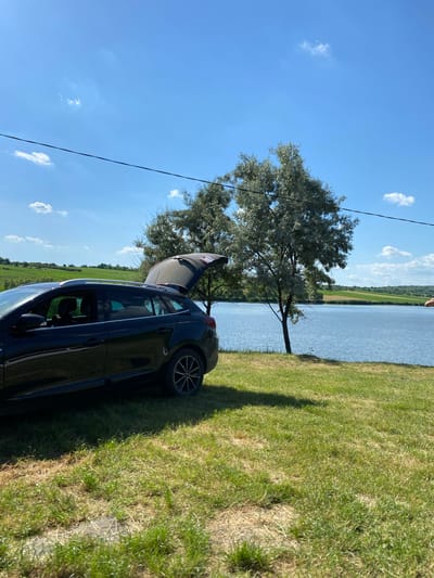 In Găleștii Noi, Moldova, on a sunny day with a bright blue sky dotted with a few white clouds, a black station wagon is parked on a grassy area next to a tranquil lake. The car's trunk is open, suggesting it might be in use for a picnic or outdoor activity. Two trees with silvery-green foliage stand between the car and the lake. The landscape beyond the lake features rolling green hills. The overall scene evokes a peaceful, natural setting ideal for recreation.