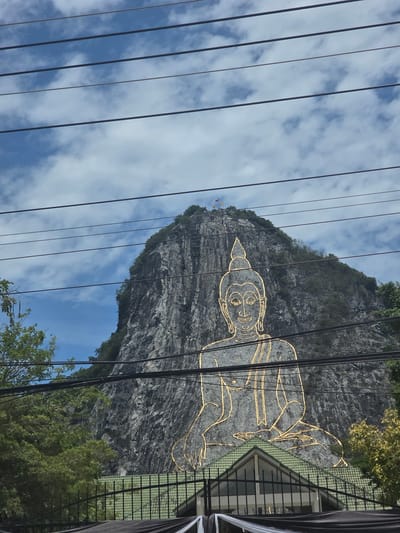 In Tambon Bang Sare, Thailand, a large Buddha image is carved into a limestone cliff face. The image, outlined in gold, depicts the Buddha in a seated meditation pose. The surrounding environment includes lush green trees and a blue sky with scattered clouds. In the foreground, there are power lines running horizontally across the image, and a green-roofed building with a black fence in front of it. The cliff face itself is rugged and textured, with vegetation growing in crevices.
