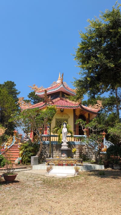 The image depicts a Buddhist temple in Lâm Sơn, Vietnam. The temple features a multi-tiered, ornate roof with decorative dragon motifs. A white statue of a deity stands in front of the temple. Vegetation surrounds the building, and a clear blue sky is visible above. The grounds appear dry and covered with foliage.