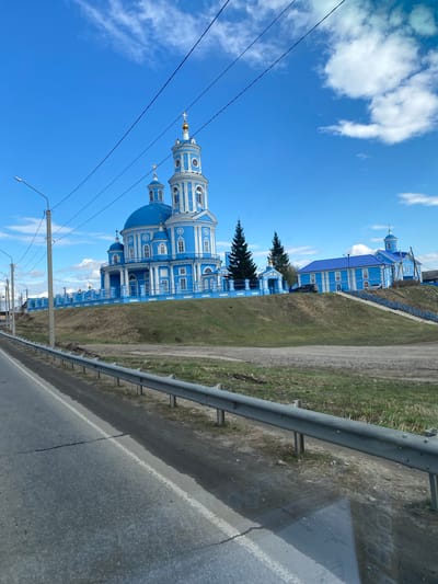 The image shows a blue and white orthodox church in Telma, Russia, captured from a roadside perspective. A grey guardrail runs along the right edge of the frame, next to an asphalt road. Beyond the guardrail, there's a strip of green grass and dirt. The church stands out with its blue domes and white architectural details, contrasting against a clear blue sky dotted with scattered white clouds. Electrical power lines run diagonally across the sky in the lower left part of the image. A secondary, smaller blue building can be seen behind and to the right of the main church building. The overall scene is captured in daylight.
