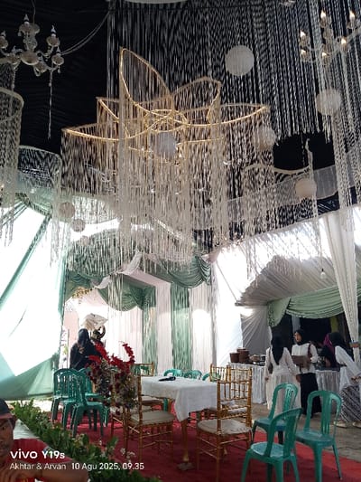 The image depicts an indoor or semi-outdoor event space in Jagapura Kulon, Indonesia, captured on August 10, 2025, at 09:35. The ceiling is predominantly dark and extensively decorated with numerous vertical white fabric tassels and multiple white spherical paper lanterns. A large, central, star-shaped light fixture with a golden frame and integrated white tassels hangs from the ceiling, along with two multi-light chandeliers visible on the left and right sides. The background features green and white fabric drapes arranged in vertical panels and horizontal swags, forming a backdrop or wall. A red carpet covers the floor.
In the mid-ground, a rectangular table draped with a white tablecloth is positioned on the red carpet. It features a floral arrangement with red and white flowers and a microphone. Around this table, several gold-colored chiavari-style chairs with white cushions are arranged. Additionally, multiple teal plastic chairs are present on the red carpet.
On the far left foreground, the head and shoulder of a person in a red shirt are partially visible, indicating the camera holder. In the mid-ground left, one individual dressed in dark clothing with a head covering stands next to a tall, white, umbrella-shaped decorative object. In the mid-ground right, three women are standing near a table holding multiple brown, lidded containers. All women wear long light-colored garments and headscarves. One woman in a white top and black skirt holds a brown bag and looks to her left. Another woman in a white top and patterned skirt looks to her right. The third woman is partially obscured. Their collective actions indicate standing and observation within the event space.