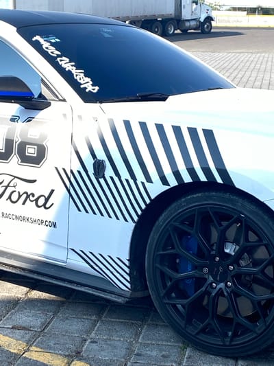 The image shows a partial view of a white Ford Mustang with black racing stripes on the side, parked on a paved surface. The car features a black rim with blue calipers and the number "86" with the word "Ford" prominently displayed, along with the website "RACCWORKSHOP.COM." A sticker on the windshield reads "RACC WORKSHOP." In the background, a truck is partially visible. The photo was taken in the city of Santa Ana Xalmimilulco, Mexico.