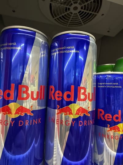 The image displays multiple cans of Red Bull energy drink, stacked horizontally inside what appears to be a refrigerator or cooler unit. The blue, silver, and red cans feature the prominent Red Bull logo, depicting two red bulls clashing against a yellow sun.
On the cans, the brand name "Red Bull" is clearly visible in red lettering, along with "ENERGY DRINK" in white. Additionally, Ukrainian text is present, stating "Енергетичний напій." (Energy drink) and "Вживати охолодженим." (Consume chilled), indicating the product's type and serving recommendation.
To the left of the cans, a white circular fan grill, typical of a refrigeration system, is partially visible, confirming the cold storage environment. The lighting suggests an indoor setting, likely a retail store, supermarket, or cafe in Donets, Ukraine, where such beverages are commonly sold and kept chilled. The cans appear new and are arranged in an orderly manner, ready for purchase or consumption.
