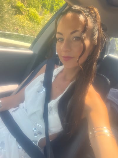 A woman is sitting in the passenger seat of a car, with her seatbelt fastened across her chest. She is wearing a white dress with a sequined pattern and ornate silver bracelets on her upper arms. Her hair is pulled back into a ponytail, with loose strands framing her face. The car appears to be moving, as indicated by the blurred background of trees and foliage, suggesting a scenic route, possibly near Đurmani, Montenegro. The sun is casting a warm glow on the scene, highlighting the woman's features and the details of her jewelry and dress.