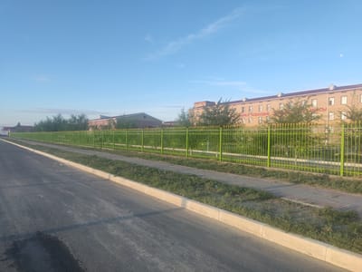 The image captures an outdoor scene in the city of Aral, Kazakhstan, on a clear day with a light blue sky and a visible moon in the distance. In the foreground, a paved road with a slight sheen runs diagonally from the bottom left to the middle of the frame. A concrete curb separates the road from a strip of grass and then a paved walkway. Beyond the walkway, a green metal fence with decorative elements encloses an area with trees and shrubs. In the background, a long, light-colored building with a dark roof is visible, featuring multiple windows and what appears to be text in red lettering on its facade. Another smaller building with a darker roof is partially visible to the left of the main building. The overall impression is one of an institutional or residential area, possibly a school or dormitory, with well-maintained grounds.