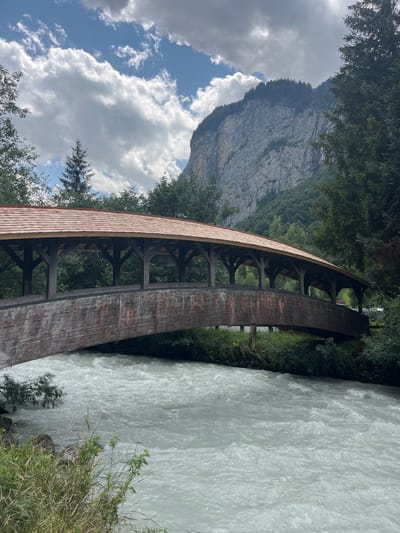 This is a full, eye-level, outdoor, vertical shot of a covered wooden bridge arching over a fast-flowing river. The bridge has a reddish-brown wooden roof and sides, with exposed wooden support beams visible underneath. The river is a milky, turbulent white, indicative of glacial meltwater, and runs from the bottom left to the middle right of the frame. The riverbanks are lined with green grass and foliage. In the background, a large, sheer, gray rock face rises dramatically, topped with a dense evergreen forest. The sky above is blue with scattered white, puffy clouds, and dappled sunlight. The scene is likely located in Wengen, Switzerland, given the characteristic mountain landscape and architecture. There are no people visible in the image.