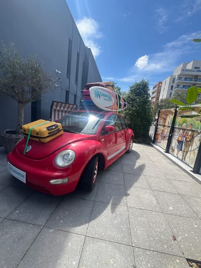 A red Volkswagen Beetle is parked on a sidewalk next to a building. The car is loaded with luggage, including two surfboards strapped to the roof and a yellow suitcase secured to the front hood. The scene appears to be a sunny day, with a clear blue sky and some scattered clouds. A tree with sparse foliage is planted in a basket next to the car. In the background, there is a modern building with a mural depicting people and a dog on its facade. A sign on the car's license plate reads "OCEANARIUM MIĘDZYZDROJE". The overall impression is of a vehicle packed for a trip, likely to the beach or a coastal destination.