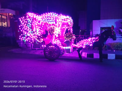 This media file depicts a horse-drawn carriage adorned with numerous bright, pink and purple lights. The carriage itself is ornate and appears to be decorated with flowers. A horse, also partially illuminated by the lights, is hitched to the front of the carriage. A person is visible in the driver's seat of the carriage, and another person's legs are seen standing near the carriage's wheels.
The scene is set at night, as indicated by the darkness of the sky and the prominent use of artificial lighting. The ground is paved, suggesting an urban or semi-urban environment, possibly a street or a decorated pathway. In the background, buildings are visible, some with architectural details illuminated by the ambient purple glow.
The overall impression is one of a festive event or celebration. The vibrant lights and decorated carriage suggest a parade, festival, or perhaps a special occasion. The atmosphere appears lively and cheerful, with the bright lights creating a magical and enchanting ambiance.
Visible text in the image includes a date and time stamp at the bottom left: "2026/07/01 20:13", followed by the location: "Kecamatan Kuningan, Indonesia". There is also partial text on a building in the upper left corner, reading "AHYU".