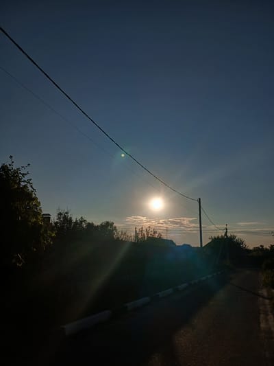 In Maslova Pristan', Russia, the image captures a rural scene with a clear blue sky at dusk. A power line stretches diagonally across the upper portion of the frame. The sun is low on the horizon, casting a bright glare and creating lens flare. Below the sky, a line of trees and houses is silhouetted against the light. A dirt road runs from the foreground into the background, marked by a curb with black and white stripes. The overall impression is of a quiet evening in a village.